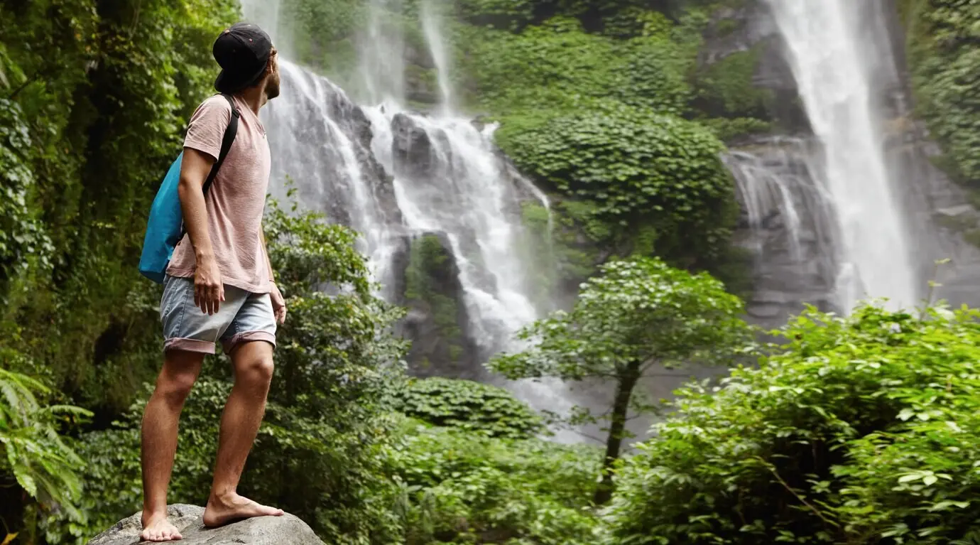 Ein junger, barfüßiger Tourist mit Baseballkappe steht auf einem großen Stein und blickt auf den hinter ihm liegenden Wasserfall in schöner exotischer Natur zurück. Der bärtige Reisende genießt die Tierwelt beim Wandern im Regenwald.